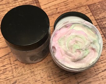 May include: Two containers of whipped bath soap. One container is open and shows a swirl of pink and green soap with a pearlescent sheen. The other container is closed and has a black lid.