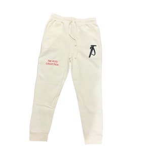 Op de afbeelding: Crèmekleurige joggingbroek met een zwart "P" logo op het rechterbeen en "THE PETTI COLLECTION" in rode tekst op het linkerbeen.