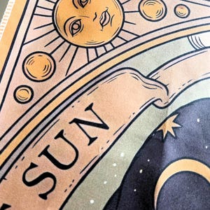 May include: A fabric tapestry featuring a stylized sun illustration with a human face, surrounded by smaller circular orbs. A banner below the sun displays the text THE SUN in black capital letters. The design includes a dark night sky background.