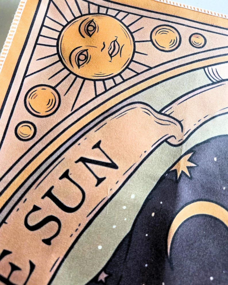 May include: A fabric tapestry featuring a stylized sun illustration with a human face, surrounded by smaller circular orbs. A banner below the sun displays the text THE SUN in black capital letters. The design includes a dark night sky background.
