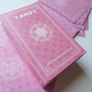 May include: A pink tarot card deck with a matching box. The box features the word "TAROT" in white, along with a floral design. The cards and box have a dotted pattern. The text "Let's share the world of Tarot" is also visible.