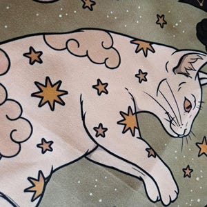 May include: A light beige cat illustration featuring gold star patterns and soft cloud shapes. The cat is depicted in a reclining position against a muted olive green background filled with small white dots and additional gold star motifs.
