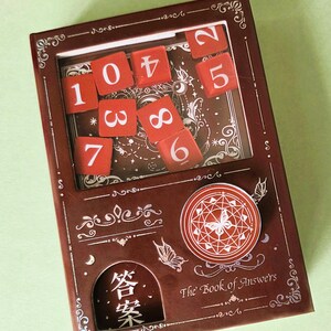 May include: A brown box with a window displaying red number tiles. The box is decorated with ornate designs, butterflies, and the text "The Book of Answers" and Chinese characters. A round sticker with a butterfly design is on the box.