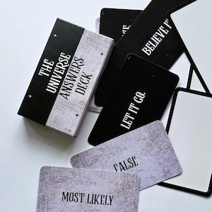 May include: A deck of cards titled "The Universe Answers Deck" with a black and white box. The cards are black with white text, including "BELIEVE IT" and "LET IT GO." Some cards have a gray, textured background with phrases like "MOST LIKELY" and "FALSE."
