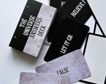 May include: A deck of cards titled The Universe Answers Deck with a black and white box. Individual cards are scattered around, featuring black backgrounds with white text like Let It Go and Believe It, or textured grey backgrounds with text.