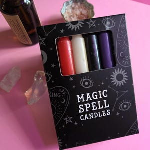 May include: A black box of "MAGIC SPELL CANDLES" with a window showing red, white, black, and purple candles. The box is decorated with celestial symbols. A bottle of liquid, a small dish of pink crystals, and clear crystals are also visible.