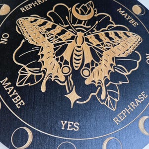 May include: A round, black wooden planchette with a laser-cut design. The planchette features a butterfly, floral elements, and the words "YES", "NO", "MAYBE", and "REPHRASE" around the edge. Crescent moon phases are also included.