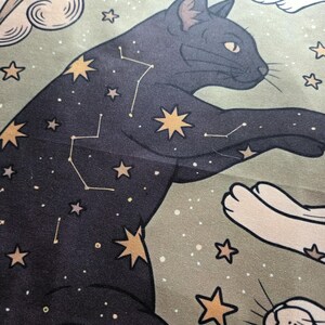 May include: A fabric print featuring a black cat adorned with gold stars and white constellation lines against a sage green background. The design includes stylized clouds and additional gold stars scattered across the surface of the material.
