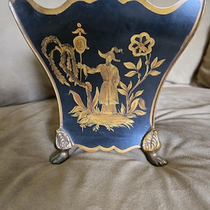 May include: A black fan-shaped decorative container with gold trim and a scalloped top edge. The front features a gold painted illustration of a figure in a hat holding a staff, flanked by a willow tree and a flower. It stands on two ornate gold feet.