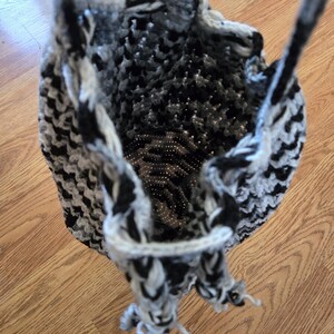 May include: A handmade crochet bag in black, white, and gray. The bag has a drawstring closure and two handles. The crochet pattern creates a textured appearance. The bag is designed to be a tote bag.