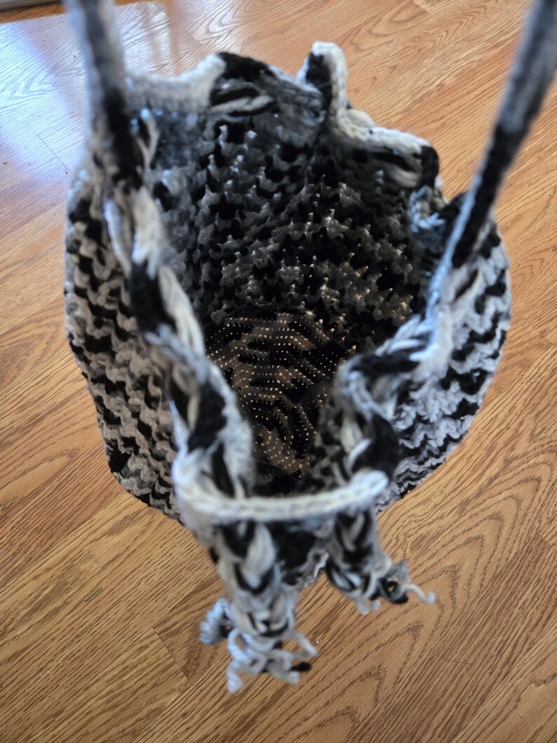 May include: A handmade crochet bag in black, white, and gray. The bag has a drawstring closure and two handles. The crochet pattern creates a textured appearance. The bag is designed to be a tote bag.