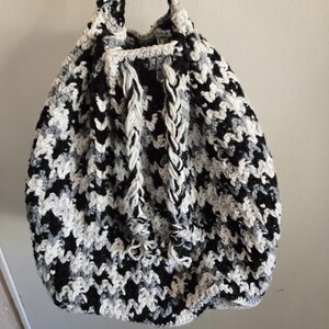 May include: A handmade crochet bag in a houndstooth pattern of black, white, and gray. The bag has a drawstring closure and braided straps. The bag is hanging against a plain white wall.