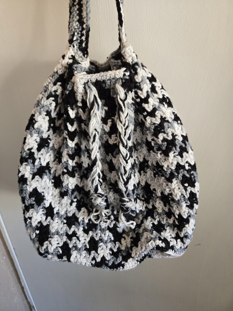 May include: A handmade crochet bag in a houndstooth pattern of black, white, and gray. The bag has a drawstring closure and braided straps. The bag is hanging against a plain white wall.