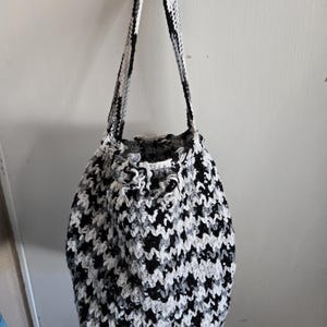 May include: A handmade crochet bag in a houndstooth pattern of black, white, and gray. The bag has a drawstring closure and long straps for carrying over the shoulder. The bag is hanging against a white wall.