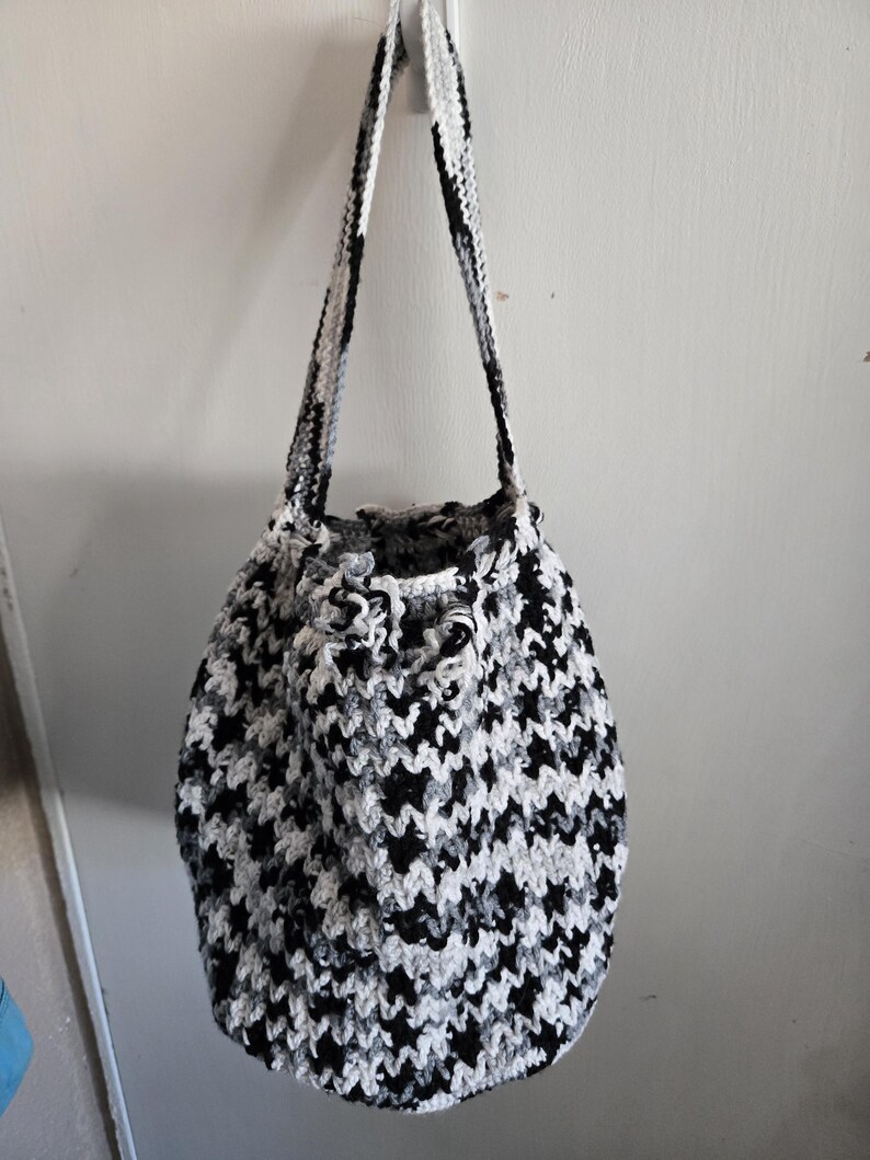 May include: A handmade crochet bag in a houndstooth pattern of black, white, and gray. The bag has a drawstring closure and long straps for carrying over the shoulder. The bag is hanging against a white wall.