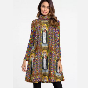 Op de afbeelding: Een tuniekjurk met lange mouwen met een glas-in-loodraamontwerp met een religieuze figuur. De jurk heeft een hoge halslijn en een kleurrijk, geometrisch patroon. De tekst "Saint Catherine of Siena" is zichtbaar op het ontwerp.