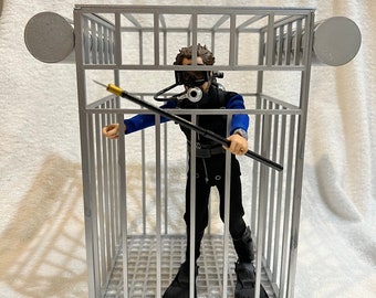 May include: A gray metal cage with a figure wearing a scuba diving suit and holding a spear inside. The cage has a metal grate floor and is open on all sides.