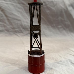 May include: A miniature buoy model with a red cylindrical base and a dark, weathered metal tower. The tower has a cross-braced design and a small red top, reminiscent of a lighthouse or nautical marker.
