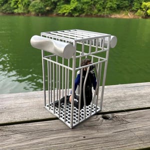 May include: A silver metal cage with a diver inside, set against a lake and trees. The cage has a rectangular top and bottom with vertical bars and cylindrical accents. The diver is wearing a black wetsuit and scuba gear.