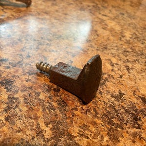 May include: A rusty metal hook with a screw thread. The hook is bent at a 90-degree angle and has a rounded tip.