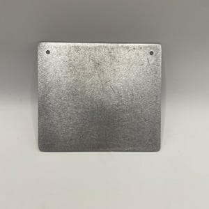 May include: A square, brushed metal plate with rounded corners. The plate has two small holes near the top edge and a textured surface with visible scratches. The metal has a silver-gray color and reflects light.