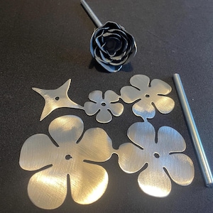 May include: A set of metal flower stencils, including a large flower with five petals, a smaller flower with four petals, a star-shaped stencil, and a metal rod. The stencils are silver and have a brushed finish.