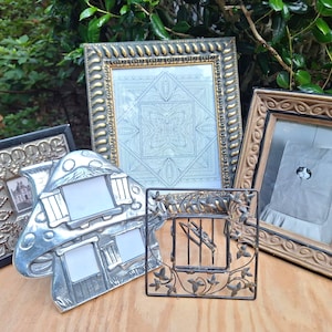 May include: A collection of four picture frames: a large rectangular frame with a gold textured border, a black frame with a silver embossed edge, a silver-toned mushroom-shaped frame with two openings, and a small square black metal frame.