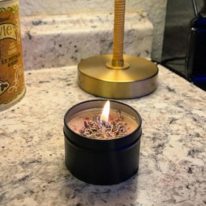 Pu&ograve; includere: Una candela in latta nera con un top di cera rosa e bianco e fiori di lavanda secchi. La candela &egrave; accesa e la fiamma &egrave; visibile.