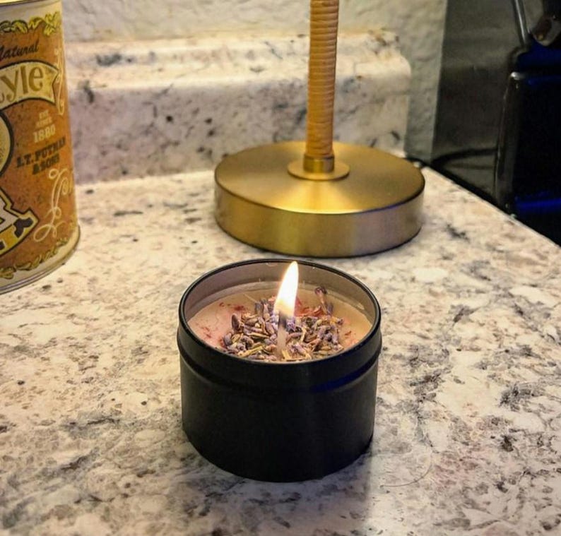 Pu&ograve; includere: Una candela in latta nera con un top di cera rosa e bianco e fiori di lavanda secchi. La candela &egrave; accesa e la fiamma &egrave; visibile.