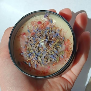 Pu&ograve; includere: Primo piano di una candela con una base di cera marrone chiaro e uno strato di fiori di lavanda secchi e cristalli rosa in cima.