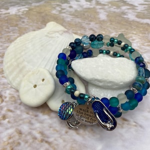 May include: A beaded bracelet with shades of blue and teal, featuring charms of a mermaid tail, a flip-flop, and a quote. The bracelet is displayed with seashells and a beach background, evoking a coastal theme.