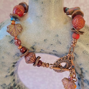 May include: A bracelet featuring a mix of textured copper-toned metal beads, round red glass beads with a dimpled surface, and leaf-shaped charms. The piece is secured with a circular toggle clasp and includes a small dangling leaf charm at the closure.