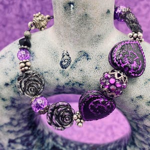 May include: A purple and black beaded bracelet with silver accents. The bracelet features various beads, including rose-shaped beads, heart-shaped beads, and faceted beads. The bracelet is displayed on a light blue and gray ceramic vase.