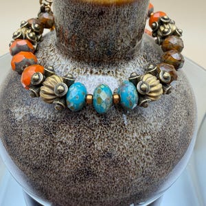 May include: A beaded bracelet featuring a mix of faceted orange beads, brown marbled beads, and three central turquoise-colored beads with a speckled pattern. The beads are separated by antique brass-toned metal spacers with a ribbed design.