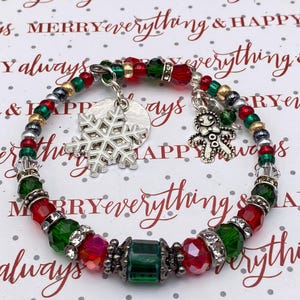 May include: A festive Christmas bracelet with red, green, and silver beads. The bracelet features snowflake and gingerbread man charms. The background has the words "Merry Everything & Happy Always" in a repeating pattern.