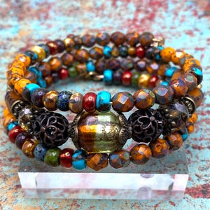 May include: A multi-strand wrap bracelet featuring a variety of faceted and smooth beads in shades of amber, turquoise, red, and metallic bronze. The design includes two dark bronze filigree spacer beads flanking a central translucent glass bead.