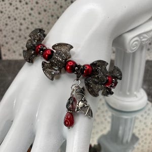 May include: A dark gray bracelet featuring bat-shaped charms with silver glitter accents, interspersed with red beads. A silver bat charm dangles from the bracelet, along with red and black teardrop beads. The bracelet is displayed on a white hand.