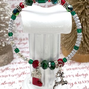 May include: A Christmas-themed bracelet with a mix of green, red, and silver beads. The bracelet features a red stocking charm and a silver reindeer charm. The background includes decorative Christmas trees and festive text.