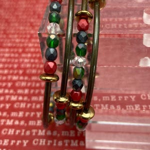 May include: Set of four beaded bracelets with a Christmas theme. Each bracelet features a series of red, green, and clear beads, separated by gold-colored metal accents. The bracelets are stacked together, creating a layered look.