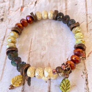 May include: A beaded bracelet featuring a mix of colors including amber, cream, and green. The bracelet has a variety of bead shapes and sizes, with leaf-shaped charms and metal accents. The bracelet is designed with an earthy, natural aesthetic.