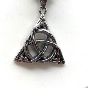 May include: A silver-colored pendant featuring a Celtic knot design, suspended from a matching chain. The pendant is a triangular shape with interwoven loops, showcasing a polished finish. The necklace is displayed against a white background.