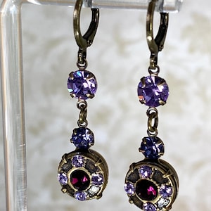 May include: A pair of dangle earrings with a vintage aesthetic. Each earring features a dark bronze-colored metal setting with purple and red crystals. The earrings are suspended from a lever-back closure.