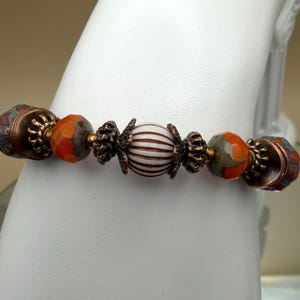 May include: A beaded bracelet featuring a central white and brown striped spherical bead flanked by copper-toned metal spacers. The design includes faceted orange and grey patterned beads, alternating with additional copper-toned metal accents.