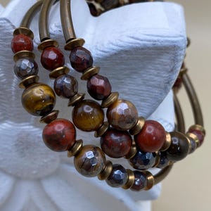 May include: Three stacked bracelets with bronze-colored metal bands and a variety of gemstone beads. The beads are in shades of red, brown, gold, and gray. The bracelets are displayed on a white decorative object.