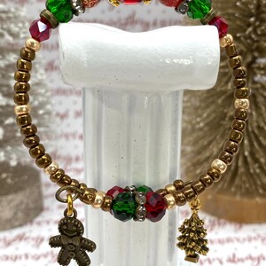 May include: A festive Christmas bracelet with red, green, and gold beads. The bracelet features a gingerbread man and Christmas tree charm. The bracelet is made of small gold beads and larger red and green faceted beads.