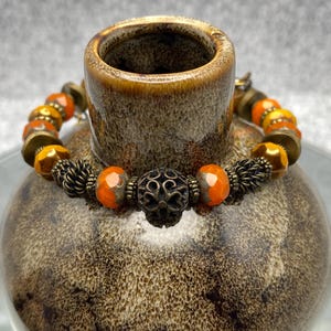 May include: A beaded bracelet with orange and yellow faceted beads, brass-colored spacers, and a decorative filigree bead. The bracelet is displayed on a brown ceramic vase with a textured surface. The bracelet is a piece of jewelry.