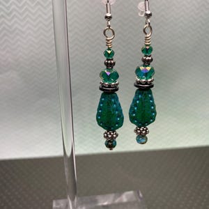 May include: A pair of dangle earrings with a teal and silver color scheme. The earrings feature a teardrop-shaped bead with a textured surface, accented by smaller beads and silver-toned metal components. The earrings are suspended from silver-colored ear wires.