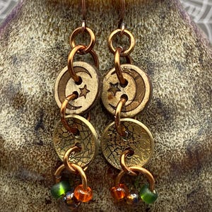 Peut inclure: Boucles d'oreilles en cuivre faites à la main, ornées de boutons en bois avec des motifs de lune et d'étoile. Les boucles d'oreilles ont un design superposé avec des anneaux en cuivre et sont ornées de perles vertes, orange et marron. Un accessoire unique de style bohème.