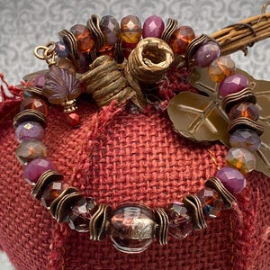 May include: A beaded bracelet featuring faceted glass beads in shades of deep red, purple, and amber, interspersed with antique bronze-toned metal spacers. A small, dark purple leaf-shaped charm hangs from the bracelet, which is circular in shape.