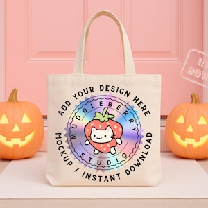 Puede incluir: Un bolso tote crema con un diseño de gato fresa y el texto "ADD YOUR DESIGN HERE". Dos calabazas talladas de color naranja flanquean la bolsa. El fondo es una puerta rosa.